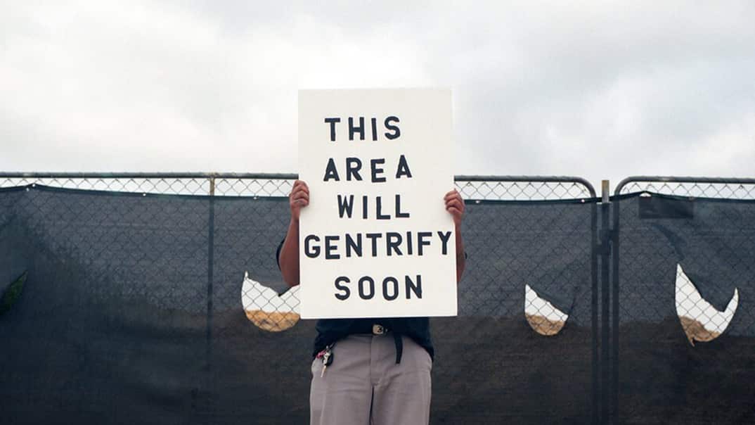 A person holds a sign in front of their face that reads "THIS AREA WILL GENTRIFY SOON"