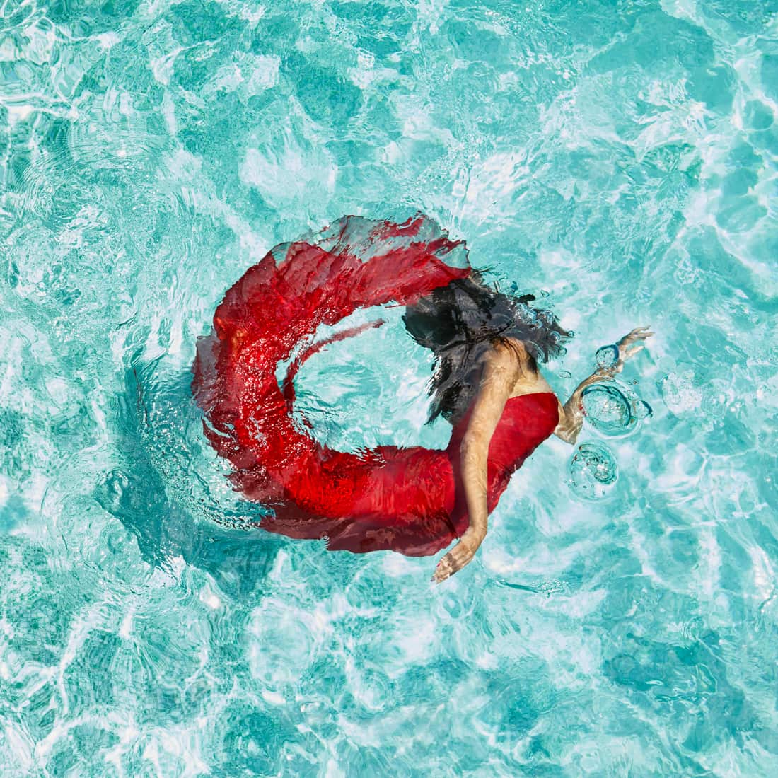 Photograph of woman in red dress swimming just below the water's surface. Her body arches back to form a circle